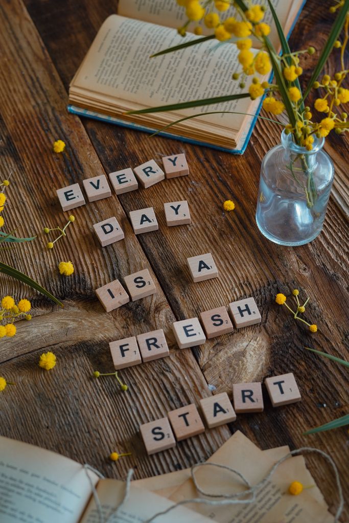 Inspirational message spelled with Scrabble tiles, surrounded by books and mimosa flowers.